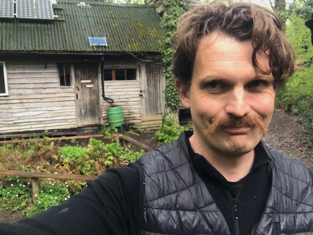 Oliver Broadbent standing smiling in front of the Longhouse, a timber building at Hazel Hill Wood to illustrate the post 'in praise of the longhouse'