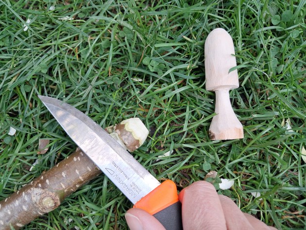 a hand in the frame holds a specialised whittling knife to a stick, with a whittled mushroom nearby
